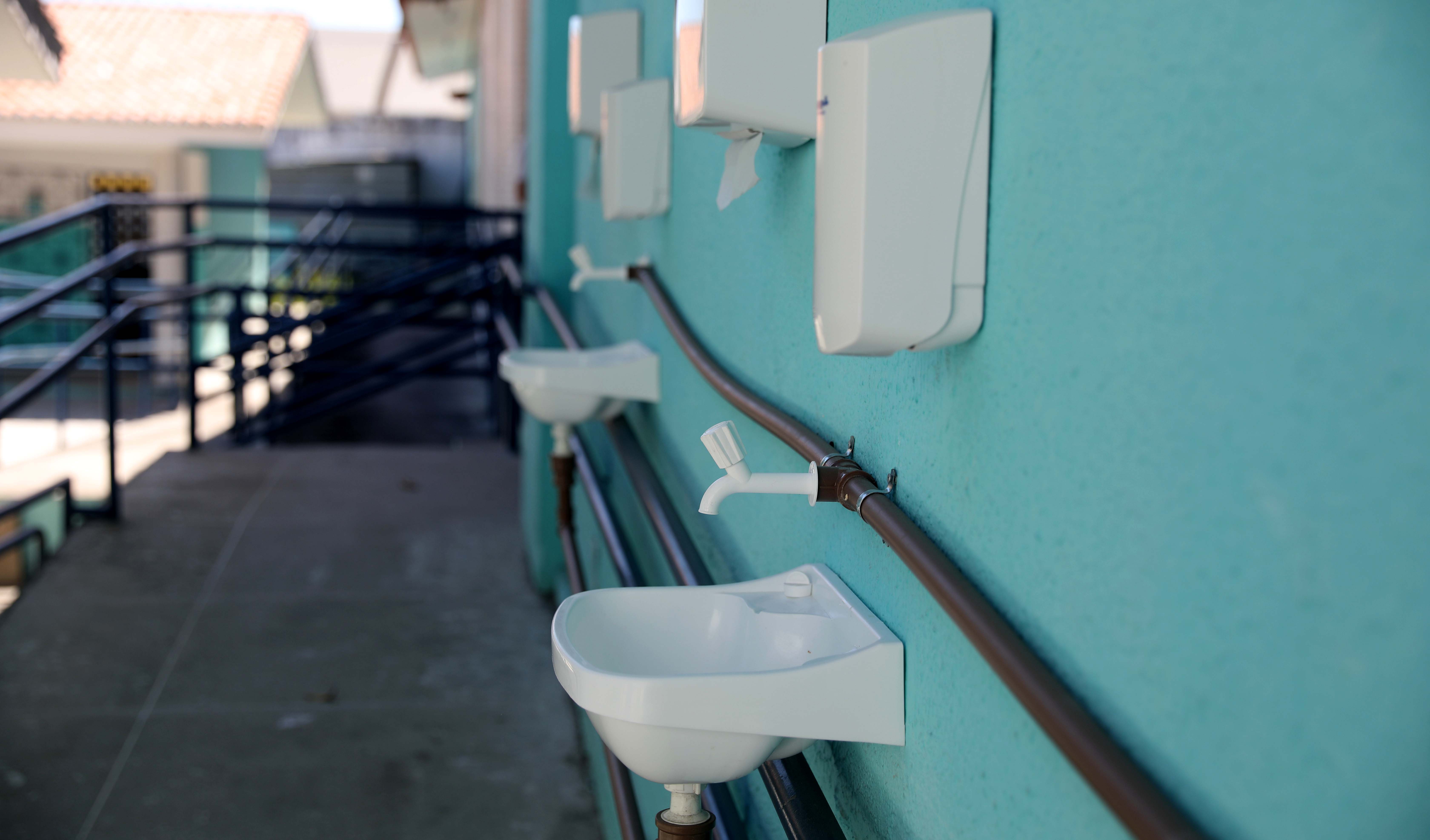 a foto mostra duas pias e recipientes de sabão líquido instaladao numa parede num pátio interno de uma escola